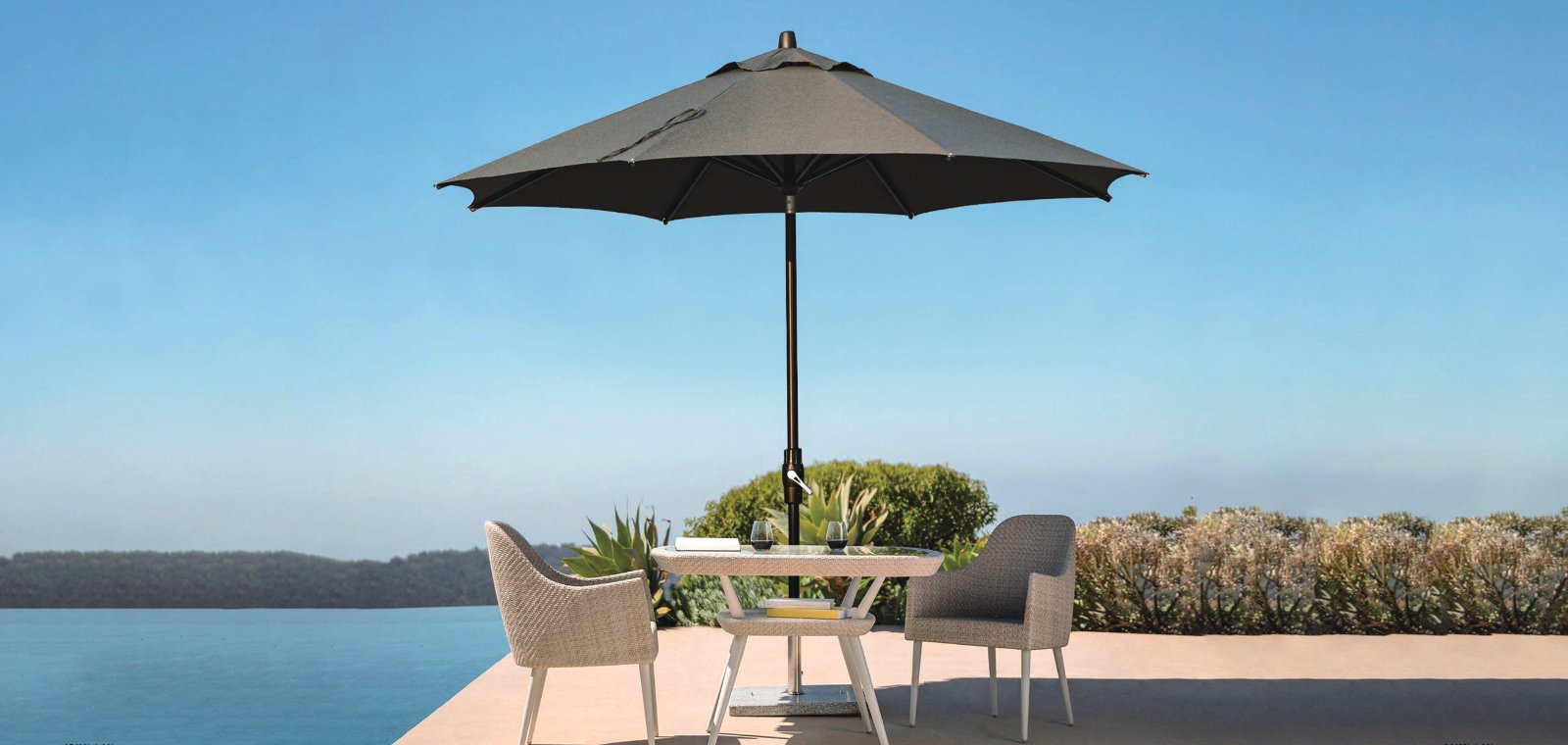 Dark gray patio umbrella shading a round woven table and two wicker chairs beside an infinity pool